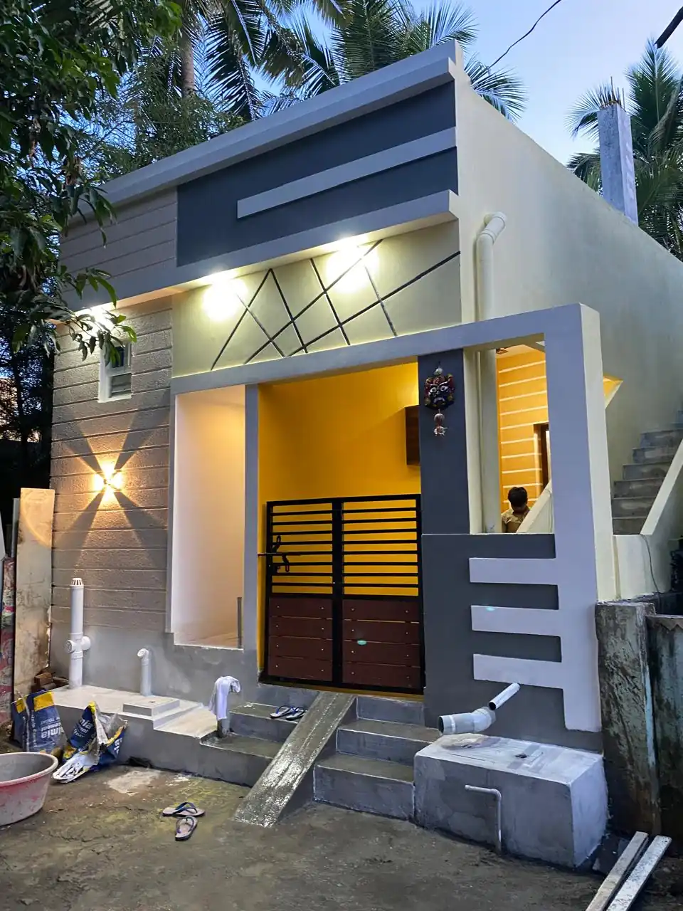 Completed single-floor house in Pammal at dusk, with a lit entrance, yellow porch wall and grey geometric façade
